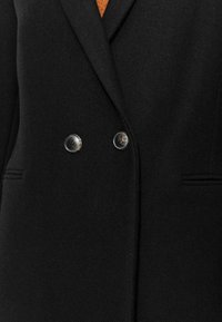 Blazer noir en tissu texturé, doté de double boutons et de poches latérales discrètes, présentant un design sur mesure.