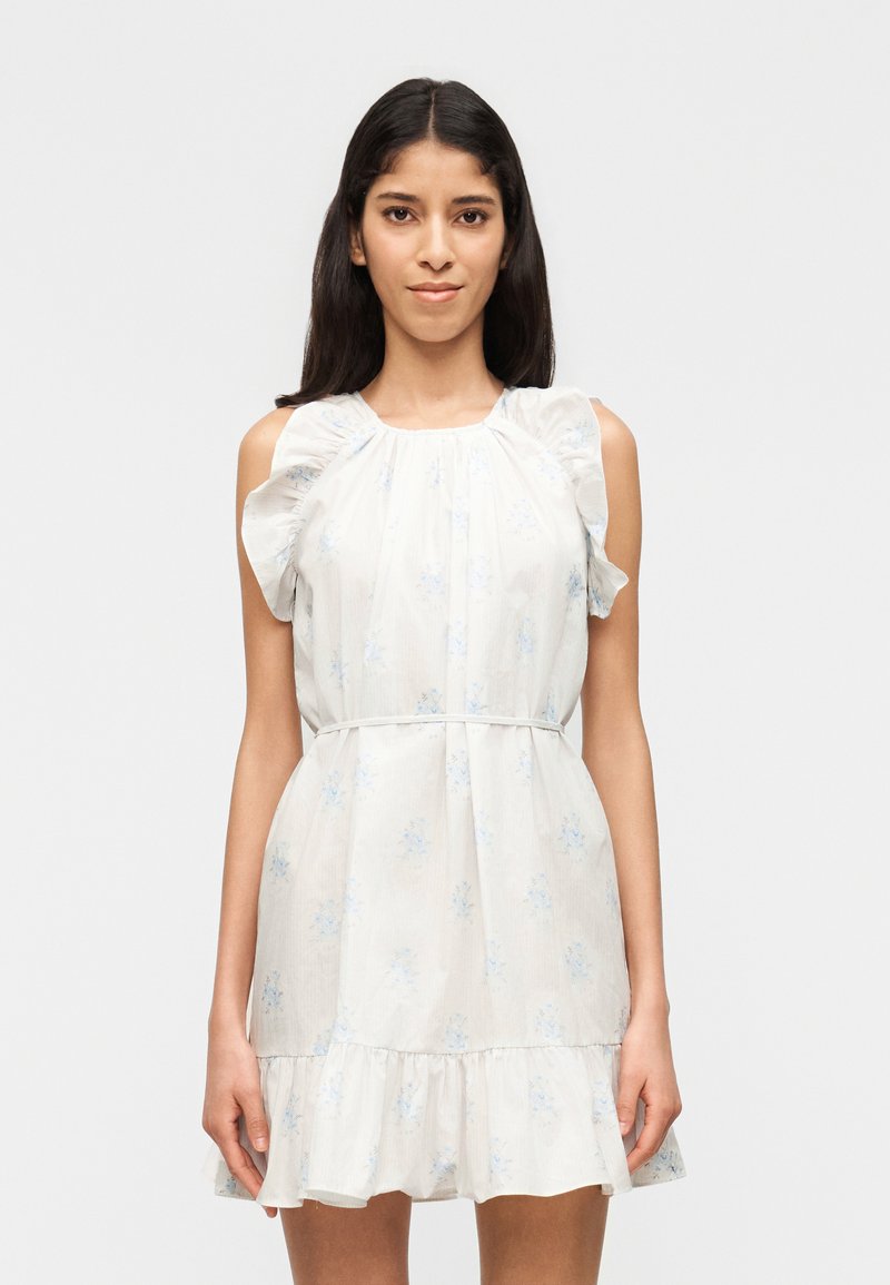 Γυναίκα με μακριά σκούρα μαλλιά, ντυμένη με λευκό αμάνικο φόρεμα με βολάν στους ώμους και μικρό μπλε λουλουδάτο μοτίβο, που στέκεται μπροστά σε ένα απλό φόντο.