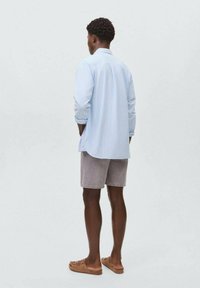 Una persona sta in piedi con le spalle alla fotocamera, indossando una camicia a righe blu chiaro, pantaloni corti grigi e sandali, su uno sfondo neutro.