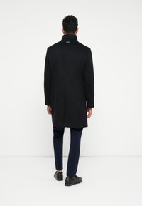 Abrigo negro con cuello alto, hecho de una tela similar a la lana. Presenta un diseño elegante, de longitud media y una abertura en la parte posterior para facilitar el movimiento. Combinado con pantalones oscuros.