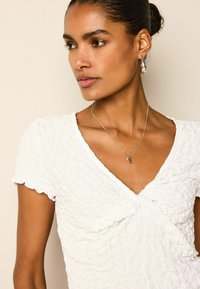 Haut blanc texturé avec un col en V, manches courtes à volants, associé à un collier en argent avec un pendentif en forme de "E" et des boucles d'oreilles en forme de goutte.