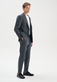 Abito grigio con rever a punta, due bottoni e tasche laterali, abbinato a una camicia bianca e scarpe nere stringate. Tessuto di trama liscia.