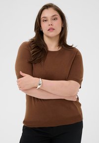 Pull à manches courtes marron en maille lisse. Comprend un col rond et une coupe décontractée. Associé à un bracelet argenté au poignet.