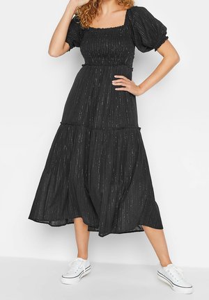 Vrouw draagt een zwarte midijurk met pofmouwen en subtiele verticale strepen, gecombineerd met witte sneakers, staand met één hand op de heup.