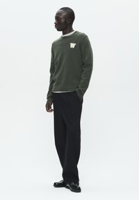 Dark green knitted sweater with ribbed cuffs and hem, featuring a white "W" logo on the chest, paired with loose black trousers and black shoes.