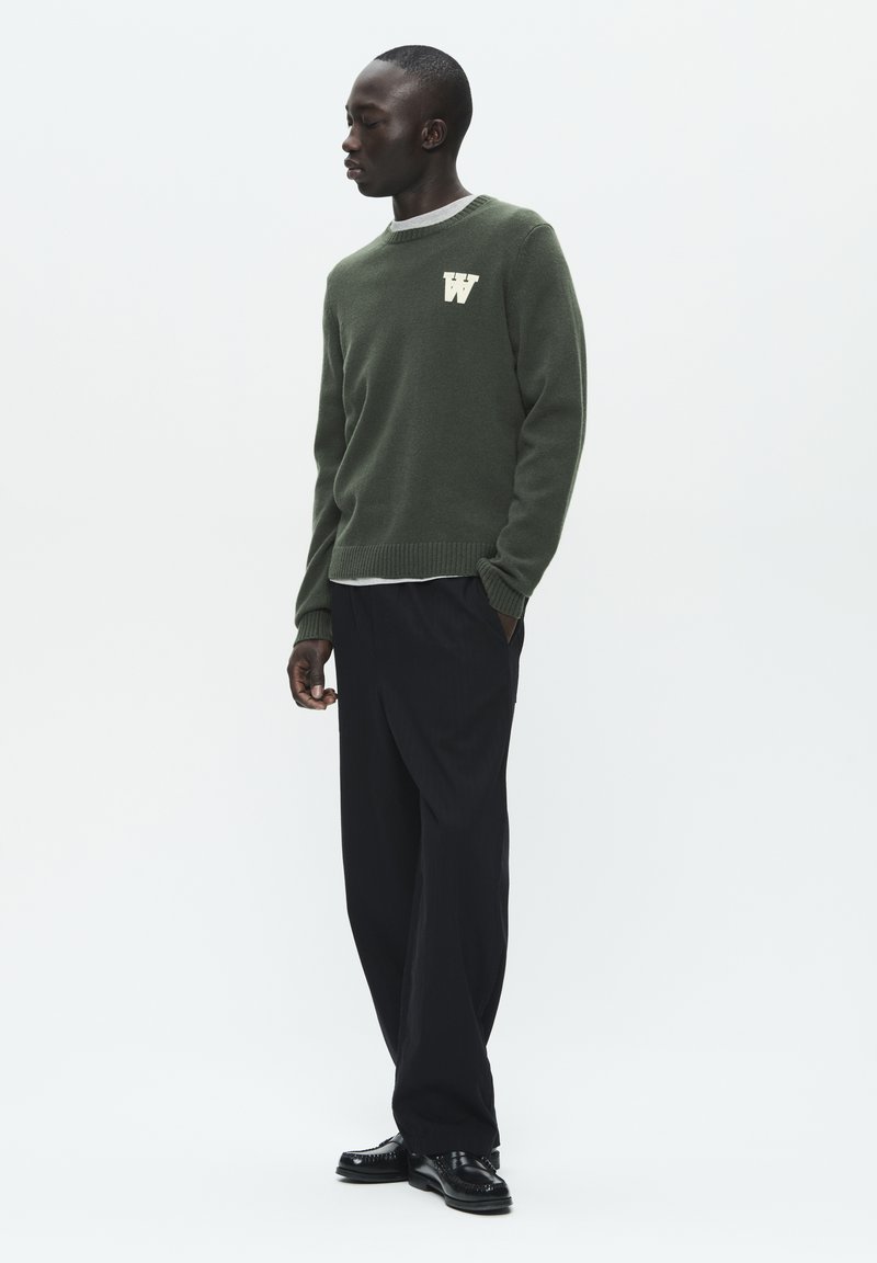 Dark green knitted sweater with ribbed cuffs and hem, featuring a white "W" logo on the chest, paired with loose black trousers and black shoes.