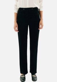 Pantalon en velours côtelé noir à cut droit, présentant deux poches avant, une fermeture boutonnée et un léger éclat. Porté avec des mocassins noirs.