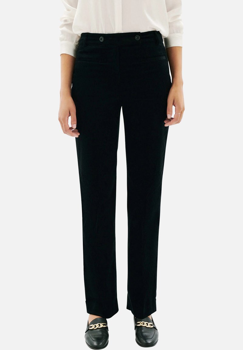 Pantalon en velours côtelé noir à cut droit, présentant deux poches avant, une fermeture boutonnée et un léger éclat. Porté avec des mocassins noirs.