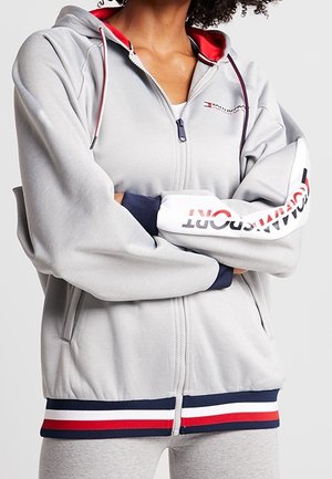 Personne portant un sweat à capuche gris zippé avec un ourlet et des manches rayés rouge, blanc et bleu, avec le logo Tommy Hilfiger.