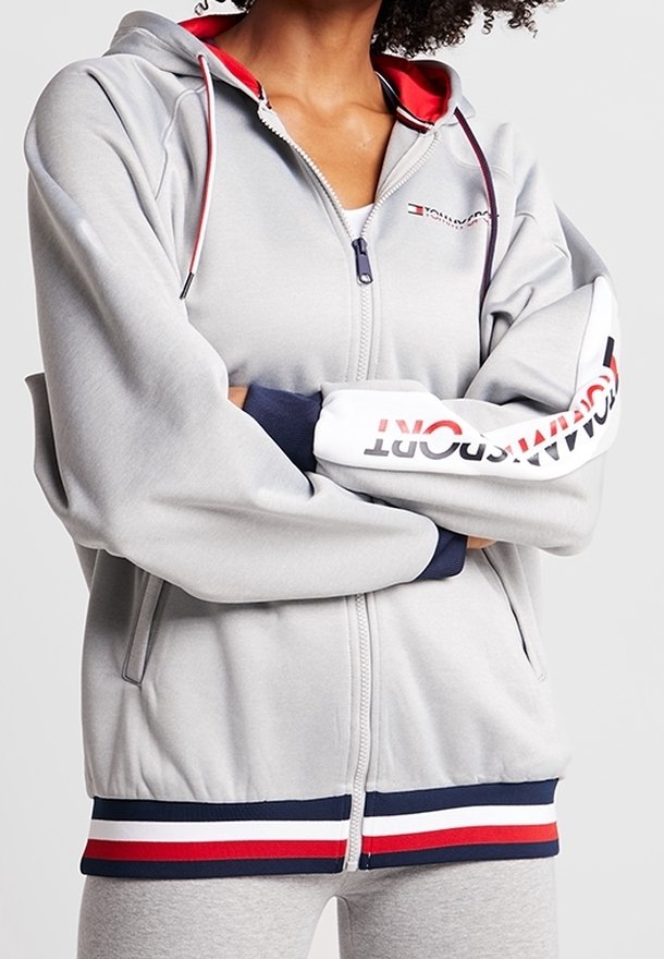 Personne portant un sweat à capuche gris zippé avec un ourlet et des manches rayés rouge, blanc et bleu, avec le logo Tommy Hilfiger.
