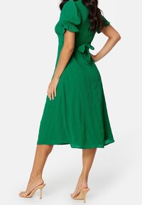 Vestido verde de largo medio con mangas abullonadas y cinturón de lazo; presenta una tela texturizada y una falda fluida. Combinado con sandalias de tacón alto en tono nude.