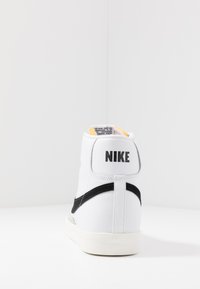 High-top Nike sneaker in white leather with a black swoosh and logo at the back. Soft yellow collar lining, rugged sole.