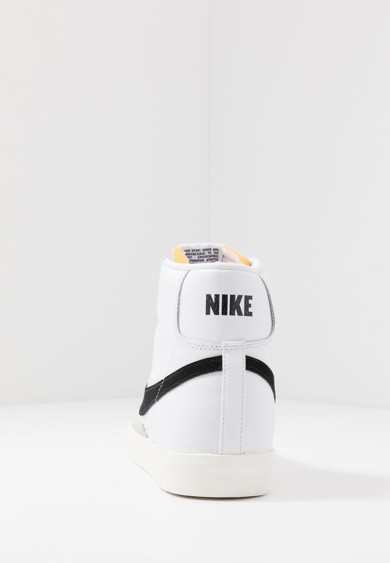 Zapatilla alta de Nike en cuero blanco con el swoosh y el logo negros en la parte trasera. Forro del collar de color amarillo suave, suela resistente.