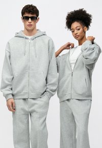 Pilkos spalvos vilkimos su užtrauktuku ir didelėmis kišenėmis, derančios pilkos spalvos laisvalaikio kelnės. Tekstūruota, minkšta medžiaga; laisvas siluetas ir nukritusios peties linijos.