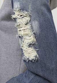Giacca di jeans con un patch logorato e bordi sfilacciati. Colore blu scuro con accenti blu chiaro. Tessuto morbido e design robusto.