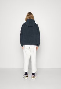 Mørkeblå fleecejakke med hætte, oversized pasform og manchetter, parret med opkrøllede hvide bukser og mønstrede sneakers.
