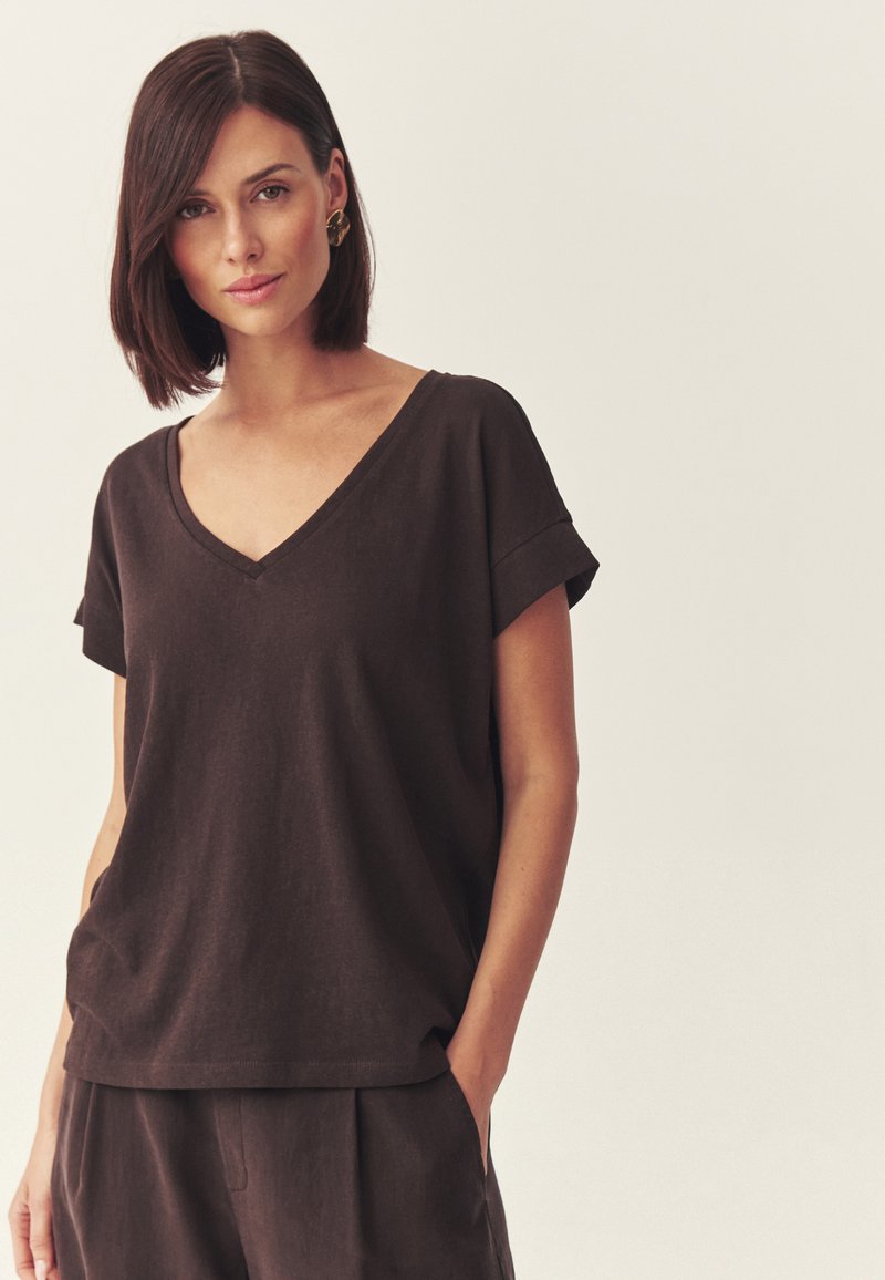 Femme aux cheveux bruns et raides porte un haut à manches courtes en V de couleur brun foncé et un pantalon assorti, debout avec une main dans la poche sur un fond clair.