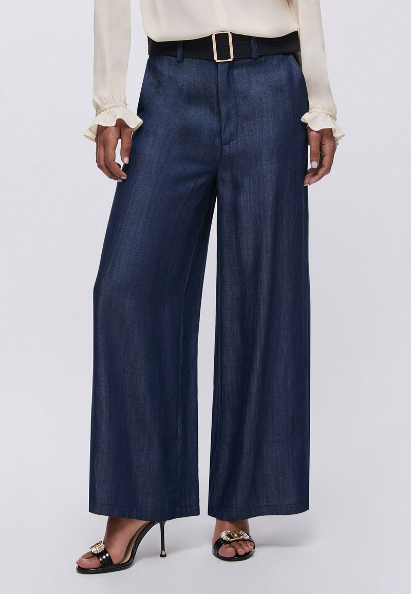 Femme portant un pantalon large bleu foncé, une ceinture noire avec une boucle dorée, un chemisier crème avec des poignets à volants et des sandales noires à talons hauts.