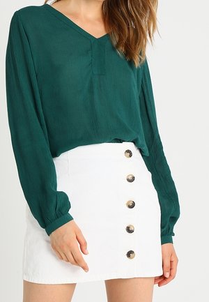 Woman wearing a green long-sleeve blouse tucked into a high-waisted white skirt with five large front buttons down the center.
