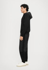 Svart hettegenser med teksturert hette, sammen med svarte joggebukser med en kontraststripe. Brukt med hvite sneakers.