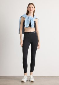 Čierne legíny s logom Calvin Klein na páse, spárované s čiernym crop topom a svetlo modrou mikinou prehodenou cez ramená.