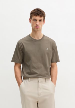 Jonge man die een bruin T-shirt met korte mouwen draagt met een klein wit logo, ingestopt in beige broek, staand met handen in de zakken.