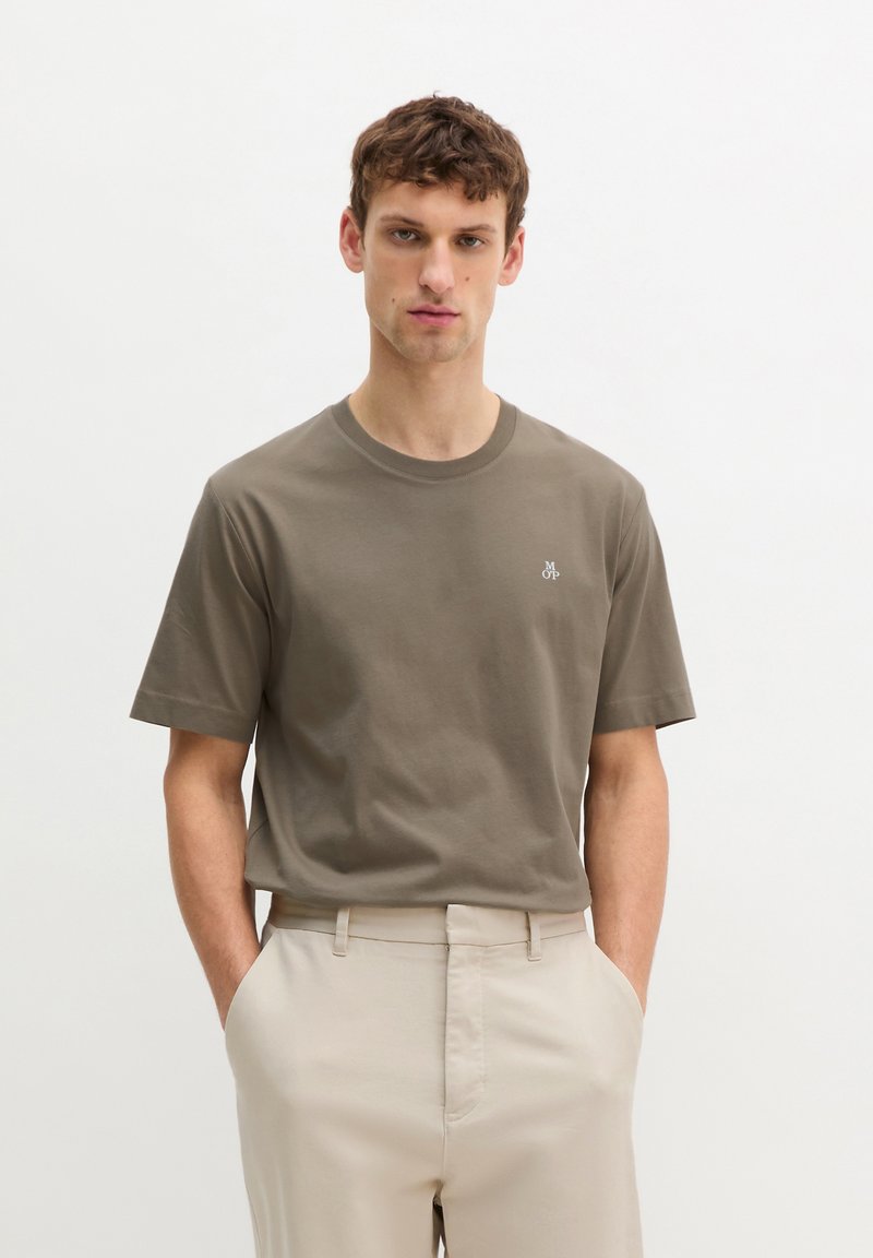 Νέος άνδρας που φοράει καφέ κοντομάνικη μπλούζα με μικρό λευκό λογότυπο, μπλουζάκι μέσα σε μπεζ παντελόνια, στέκεται με τα χέρια στις τσέπες.