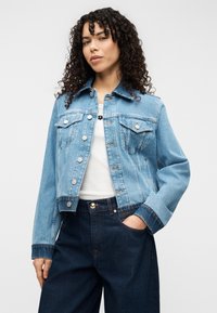 Ανοιχτό μπλε τζιν μπουφάν με κλασικό γιακά, δύο μπροστινές τσέπες και ασημένια κουμπιά τύπου snap. Φορεμένο με λευκό τοπ και σκούρα μπλε τζιν παντελόνι.