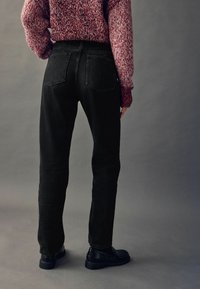 Schwarze Jeans im geraden Schnitt mit zwei Gesäßtaschen und hoher Taille, kombiniert mit einem strukturierten roten Strickpullover und schwarzen Schuhen.