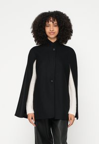 Μαύρη κάπα με ψηλό γιακά και κούμπωμα με κουμπιά, με λεία υφή τύπου μαλλιού και μανίκια μπεζ πουλόβερ σε αντίθεση.