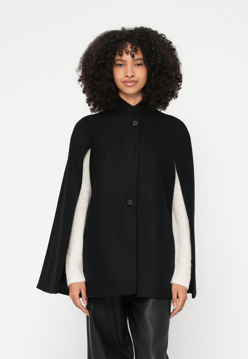 Μαύρη κάπα με ψηλό γιακά και κούμπωμα με κουμπιά, με λεία υφή τύπου μαλλιού και μανίκια μπεζ πουλόβερ σε αντίθεση.