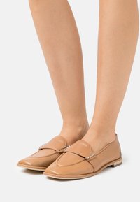 Mocassins en cuir brun clair avec un bout pointu, une sangle décorative ornée de petits clous, et une semelle plate. Texture lisse et design minimal.