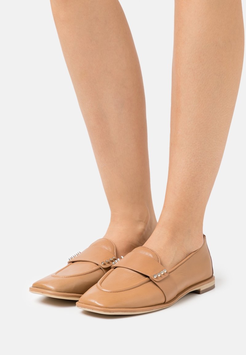 Mocassins en cuir brun clair avec un bout pointu, une sangle décorative ornée de petits clous, et une semelle plate. Texture lisse et design minimal.