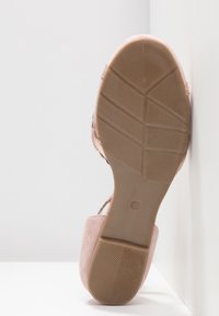 Chaussure en daim rose pâle avec une semelle en caoutchouc texturée, présentant un design épuré et un dos ouvert, inclinée contre un fond clair.