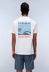 Bílá bavlněná tričko s modrou a světle modrou grafikou, obsahující vlnící se čáry a slovo "geographic" v oranžové barvě na zadní straně.