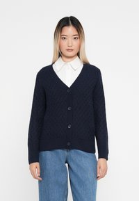 Cardigan de malha azul-marinho com um padrão de diamante, decote em V, fecho de botões e punhos canelados. Usado sobre uma camisa branca de colarinho e combinado com jeans azuis.