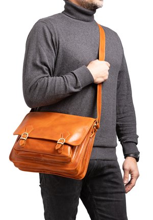 Man draagt een grijze coltrui en zwarte broek, en draagt een grote lichtbruine leren schoudertas met gespen over zijn schouder.