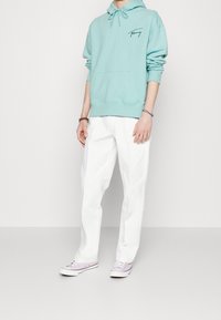 Sudadera verde menta con bolsillo frontal y cordones, combinada con pantalones blancos y zapatillas lilas claras. Diseño simple y casual.
