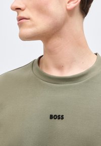 Νέος άνδρας με φανέλα σε στυλ crewneck ελιάς με μαύρο λογότυπο "BOSS" στο κέντρο του στήθους, στραμμένος αριστερά μπροστά σε απλό φωτεινό φόντο.