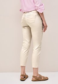 Pantalones beige de corte slim con cinco bolsillos, largos hasta el tobillo y con costuras en contraste. Llevados con sandalias marrones, con correas ajustables.