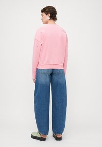 Felpa di cotone rosa con maniche lunghe, caratterizzata da piccole decorazioni. Abbinata a jeans blu larghi e scarpe verde chiaro.