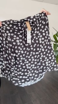 Juoda sijonas su baltu gėlių raštu laikomas rankomis viduje, matomi prisegti kainų ženklai, fone augalas ir medinė grindys.