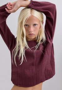 Top largo de manga larga texturizado en burdeos, con escote redondo y puños fruncidos, que presenta patrones de rayas verticales y un corte suelto.