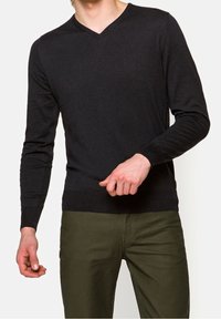 Suéter negro de cuello en V con mangas largas, hecho de tela suave; posicionado contra un fondo claro, combinado con pantalones verde oscuro.