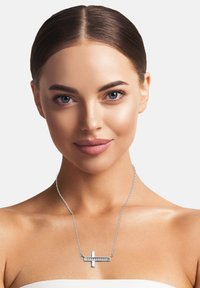 Collana d'argento con pendente a forma di croce dal design orizzontale e una fila di pietre trasparenti lungo un bordo, esposta sulla pelle nuda.