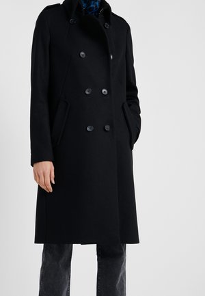 Zwarte dubbelgeknoopte lange jas met knopen, gedragen over donkere jeans, linkerhand in de zak, tegen een effen lichte achtergrond.