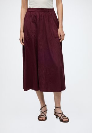 Femme portant une jupe midi bordeaux taille haute avec poches avant et des sandales noires à brides, sur un fond uni.