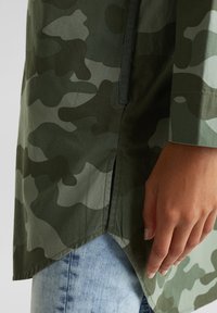 Mano descansando al lado, vistiendo una camisa de patrón de camuflaje en verde y verde oscuro con una abertura lateral, combinada con jeans de mezclilla azul claro.