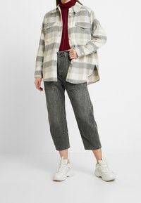 Gecontroleerde flanellen shirt in crème en grijs, met twee borstzakken. Draagt over een bordeauxrode coltrui met donkere, afgeknipte jeans en witte sneakers.