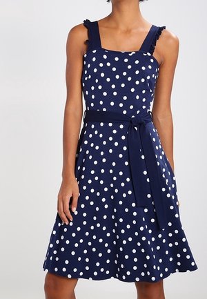Marineblauwe mouwloze jurk met witte polkadots, gerimpelde schouderbandjes en een gestrikte tailleceintuur, gedragen door een persoon van nek tot knieën.